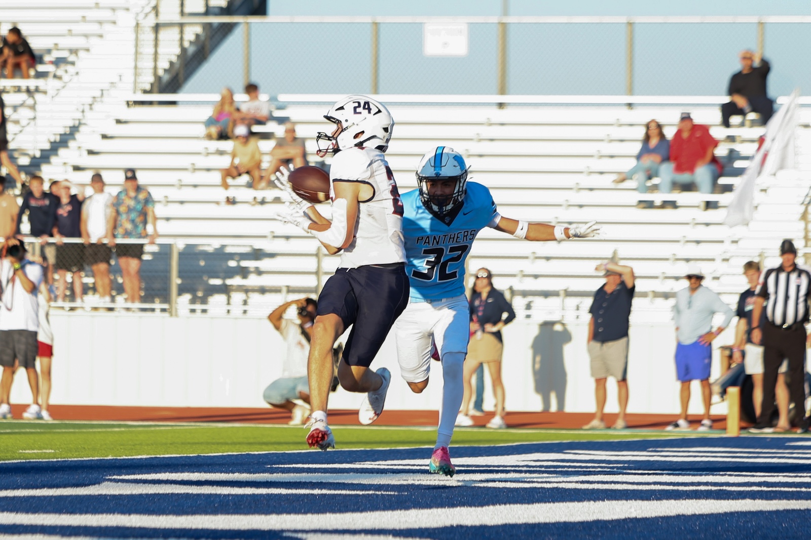 Football game: white-uniform receiver jumps with ball while pursuing end zone, pursued by a blue Panthers defender on a blue end zone turf, spectators in stands in background.