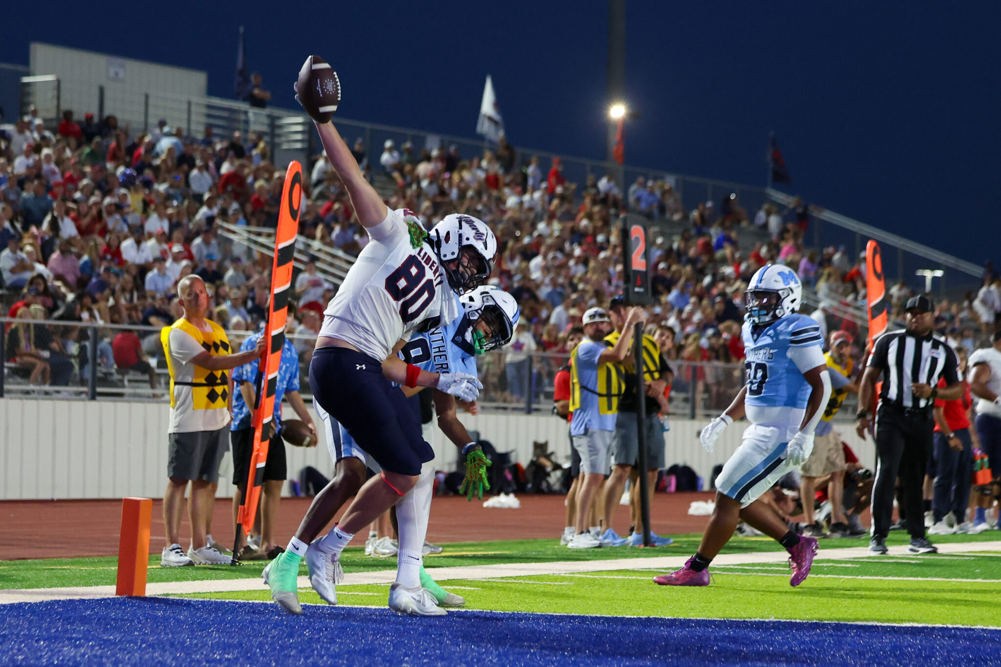 Player in white jersey reaches for a football over the goal line as teammates cheer in a blue end zone while a crowded stands watches at night.