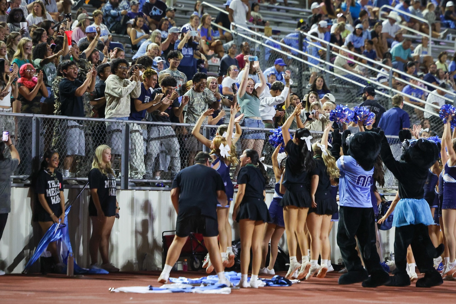 Cheerleaders on the sideline facing a cheering crowd behind a chain-link fence at a stadium rallying with arms raised and pom-poms visible.