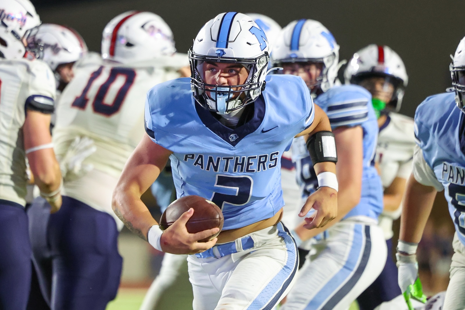 Football player in light blue Panthers uniform running with ball during a rally, defenders in white jerseys behind him.