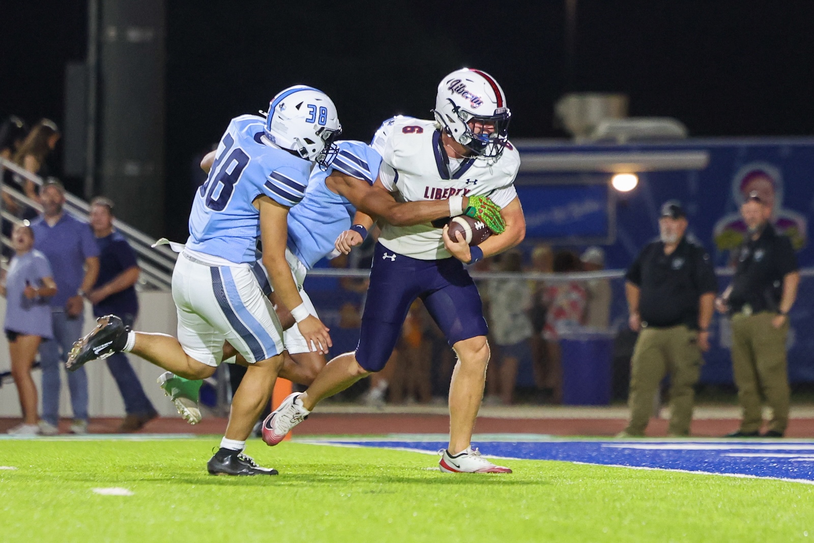 Liberty football player in white jersey holds the ball while blue-clad defenders close in during a night game on a bright green field