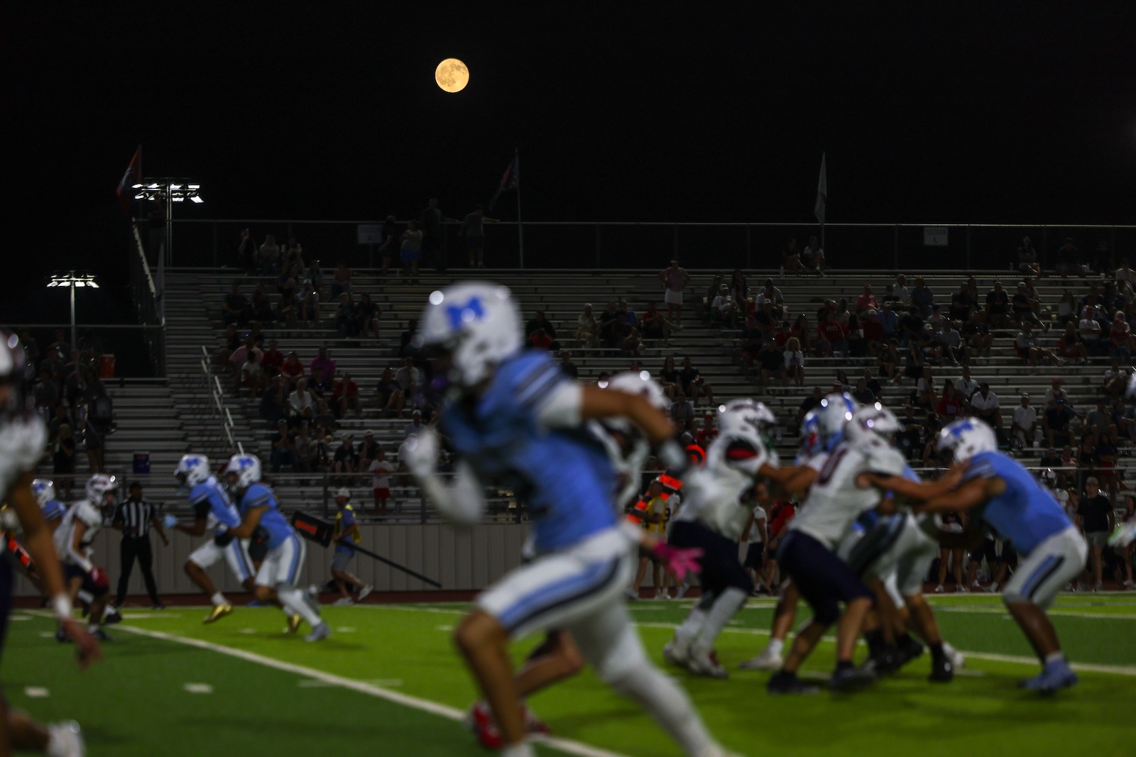 Night football game on a bright green field; players in blue and white rush as a full moon rises over the stands with spectators.
