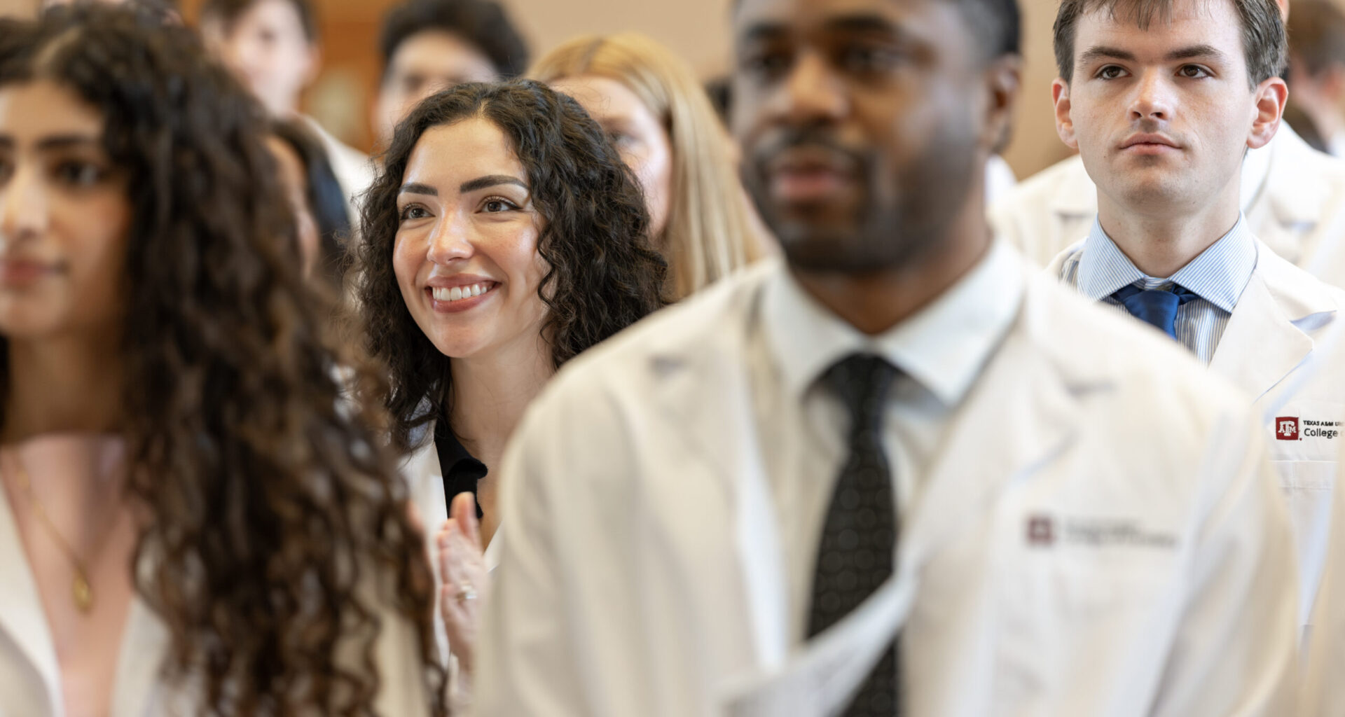 A female dental student in a lab coat watches onward while standing with other students in the audience