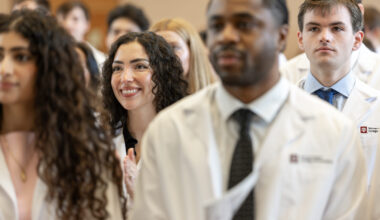 A female dental student in a lab coat watches onward while standing with other students in the audience
