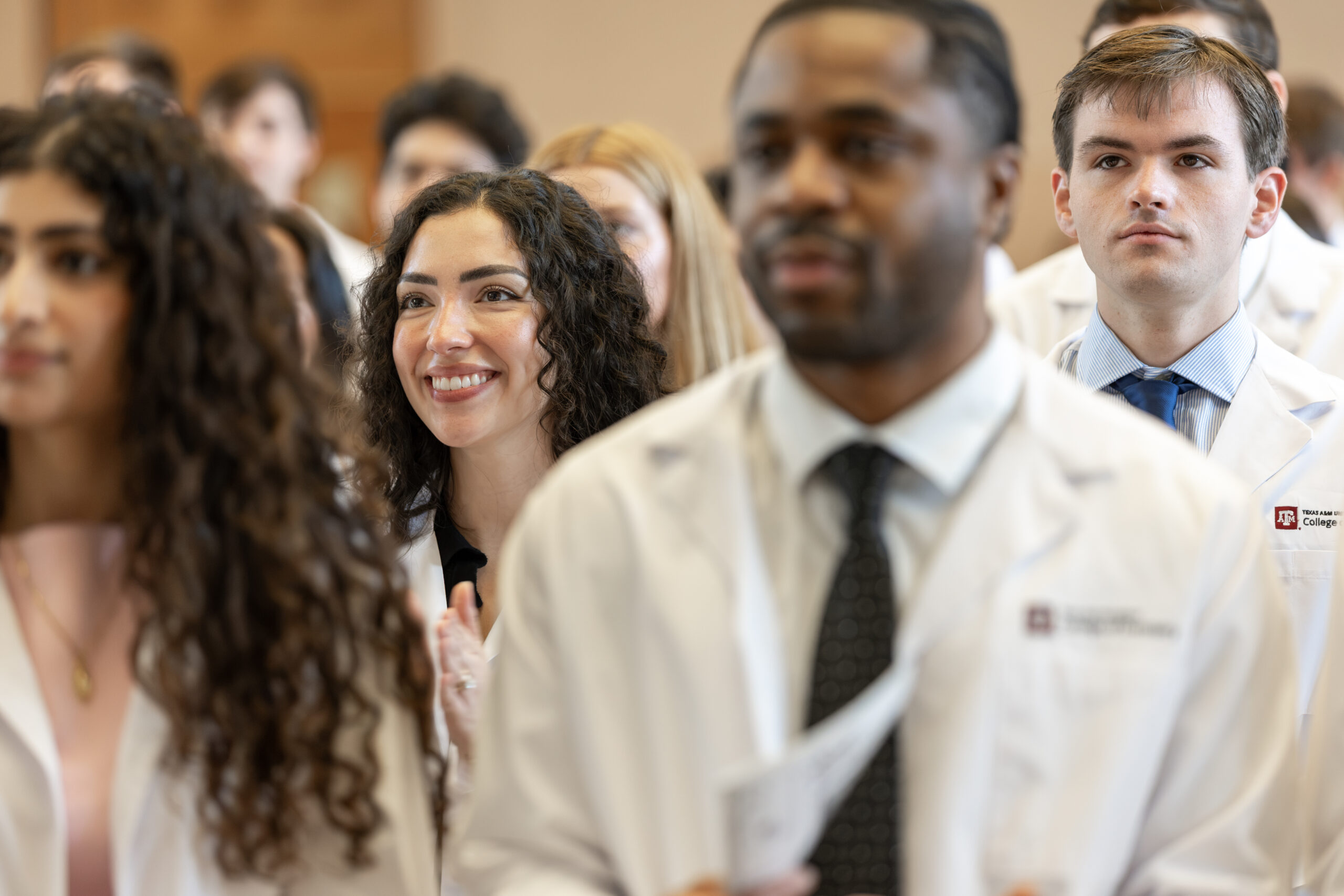 A female dental student in a lab coat watches onward while standing with other students in the audience