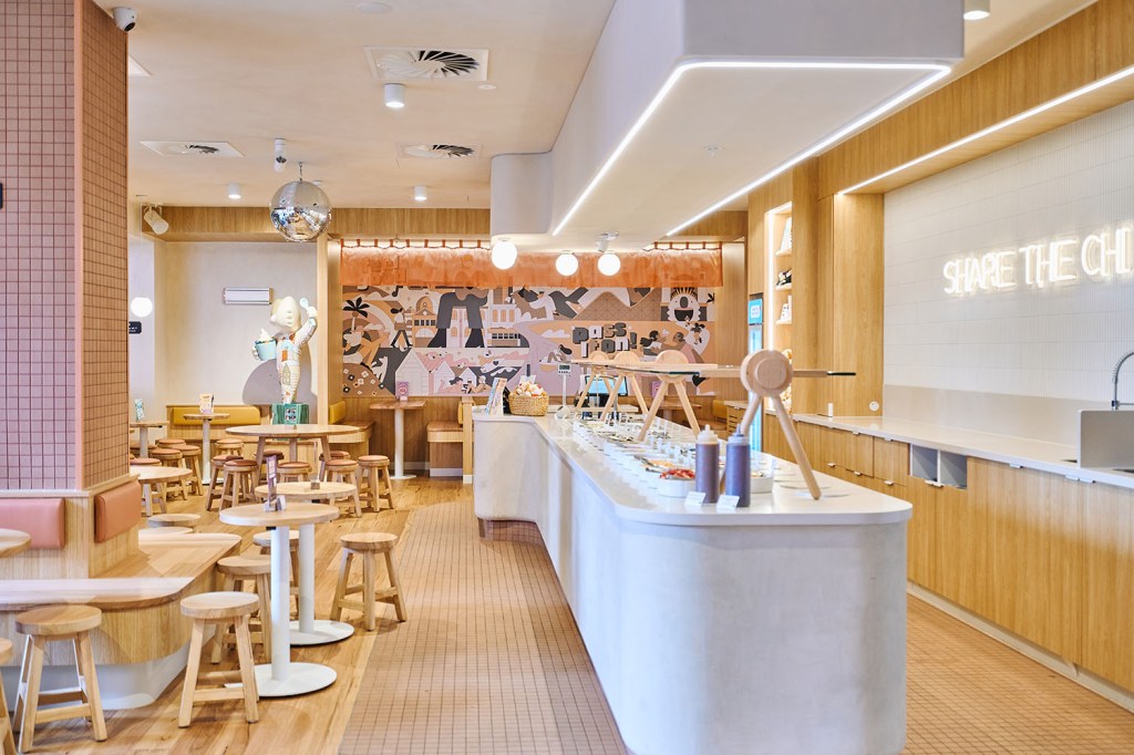 Yo-Chi frozen yogurt shop interior with pink tiled walls, wooden stools and tables, a colorful mural, disco ball, and a "SHARE THE CHI" neon sign above the self-serve yogurt counter.