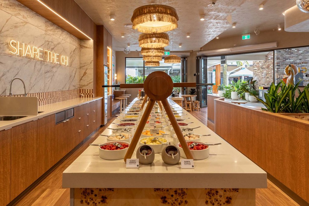 Long self-serve topping bar at Yo-Chi with bowls of fresh fruit, candy, and rainbow sprinkles under woven pendant lights, with a neon "SHARE THE CHI" sign in the background.