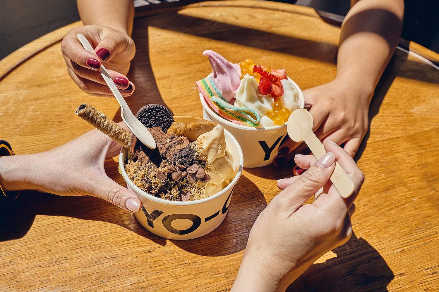 Two hands holding wooden spoons over Yo-Chi frozen yogurt cups topped with Oreo cookies, chocolate chips, wafer rolls, fresh strawberries, sour candy strips, and popping boba.