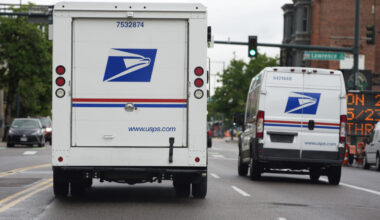 FILE - A USPS logo adorns the back doors of United States Postal Service delivery vehicles as they proceed westbound along 20th Street from Stout Street and the main post office in downtown Denver, Wednesday, June 1, 2022. USPS plans to substantially increase the number of electric-powered vehicles it