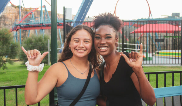 Two female students do the "Birds Up" hand sign in front of a roller coaster at Six Flags Fiesta Texas.