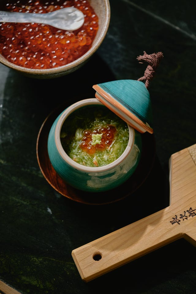 A teal ceramic pot with green broth and fish roe, beside a bowl of fish roe and a wooden serving paddle.
