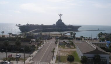 The long grey flight deck of the aircraft carrier USS Lexington moored beyond a palm lined boulevard on North Beach in Corpus Christi, under a blue sky