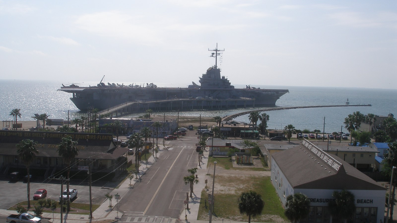 The long grey flight deck of the aircraft carrier USS Lexington moored beyond a palm lined boulevard on North Beach in Corpus Christi, under a blue sky