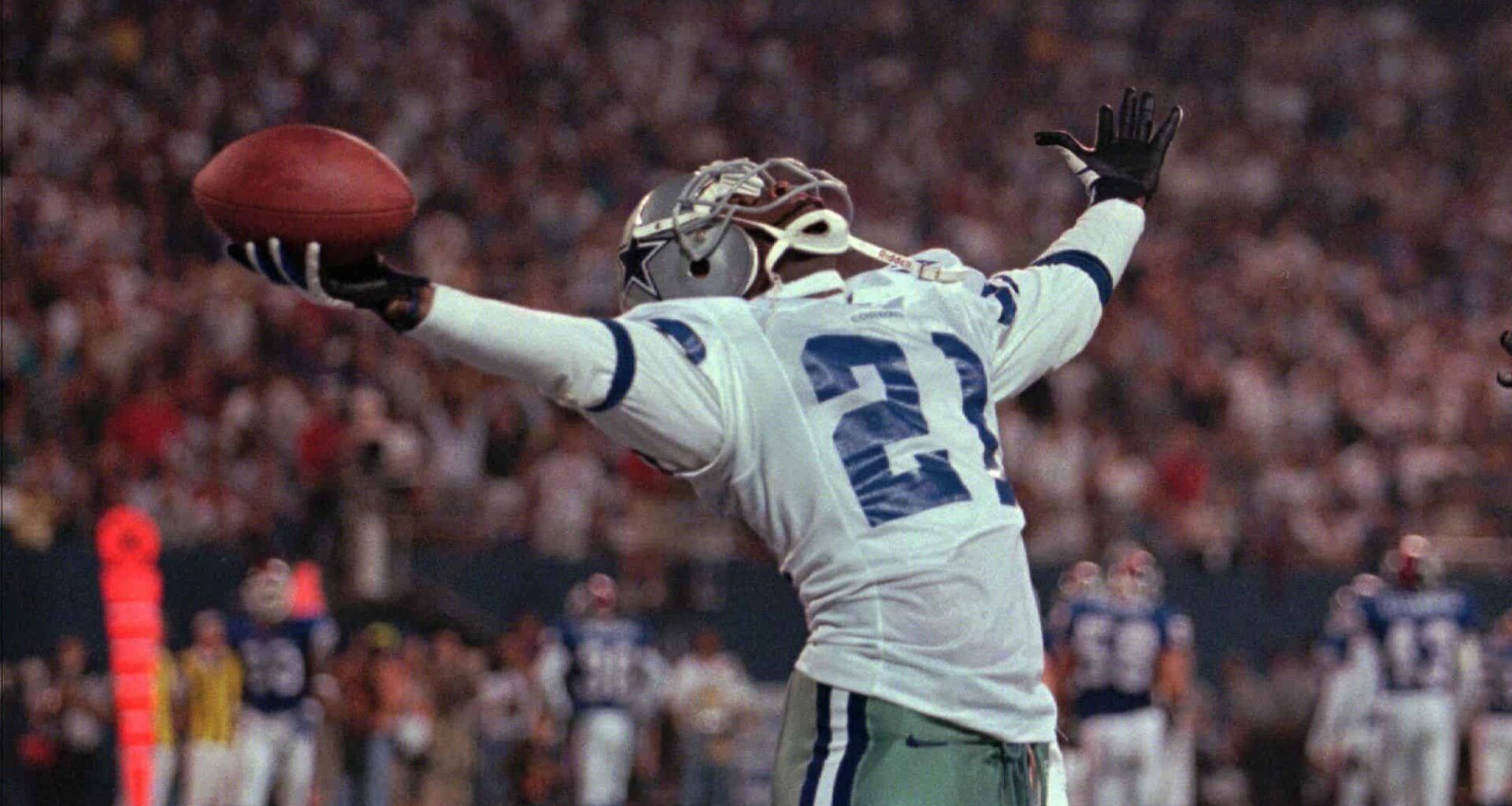 Deion Sanders celebrates while holding the football high in one hand during a Dallas Cowboys game.