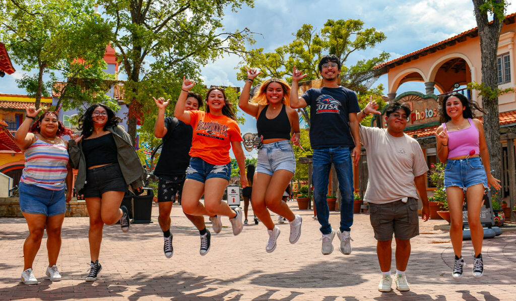 Eight young adults jump joyfully in a sunny plaza, surrounded by trees and colorful Spanish-style buildings.