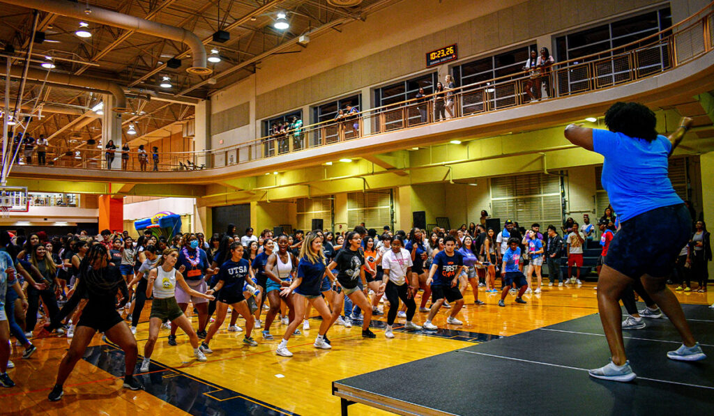 An instructor leads a large, energetic group dance workout in a crowded gym, with participants mirroring her moves and spectators watching above.