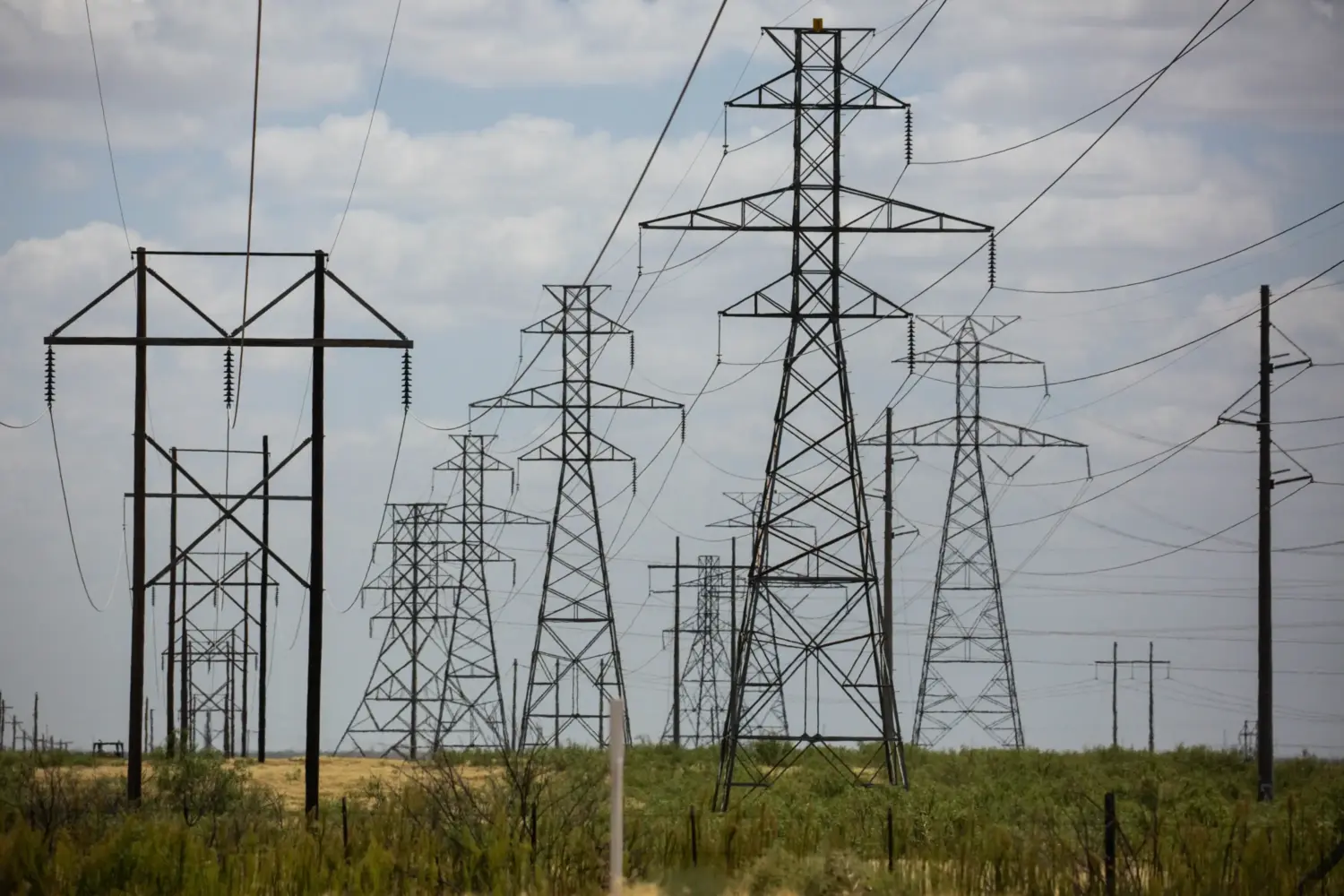 Several towers carrying electrical transmission lines crowd a swath of rural land.