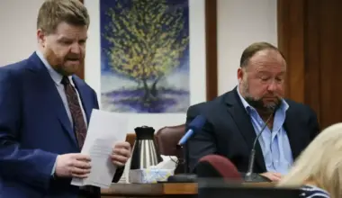 A man holding a piece of paper speaks as another man sits in a court room to answer his questions.