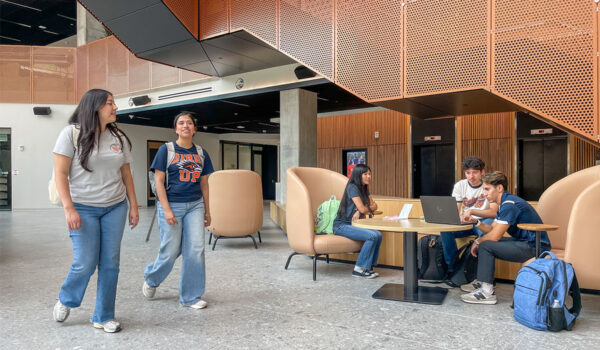 Two students walk through the San Pedro II atrium while three other students gather around a laptop on a nearby table.