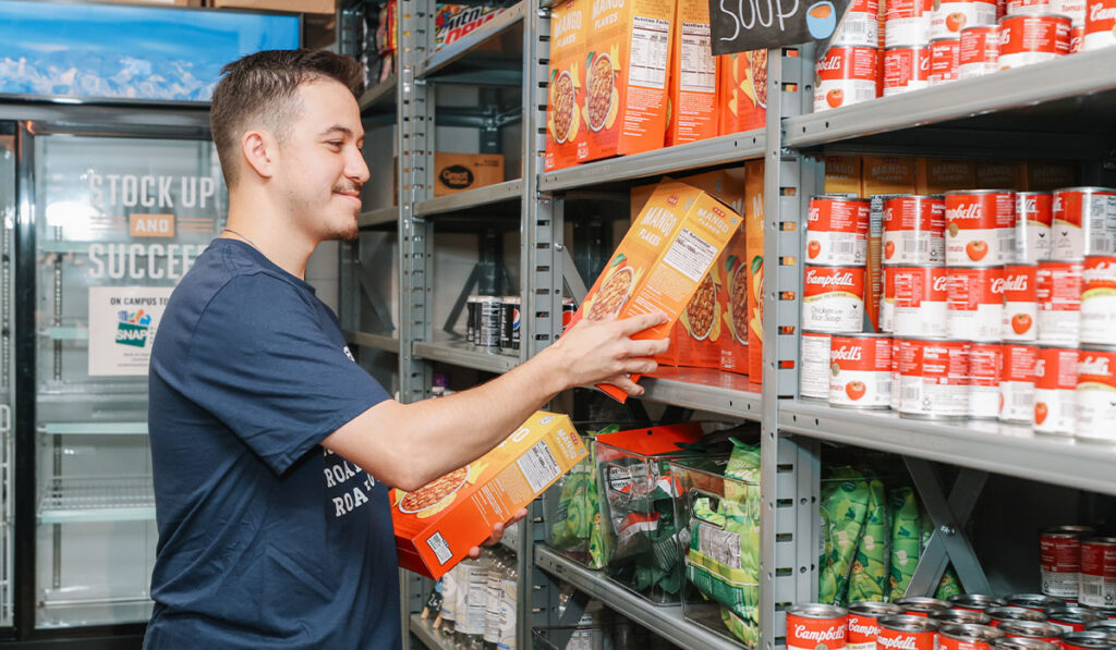 A person in a blue shirt holds two boxes of cereal, placing one of the boxes on a metal shelf with canned goods and other foods.
