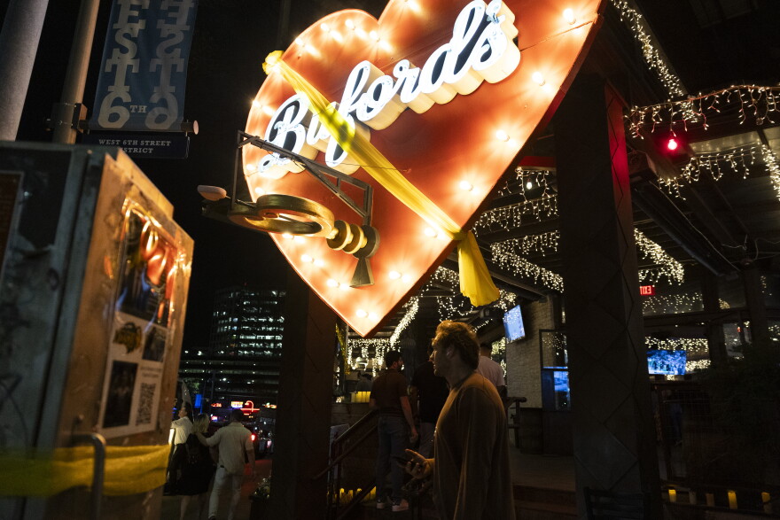 Pedestrians walk under a large lit up red heart with the text "Buford's" at night. Across the heart a yellow ribbon is tied.