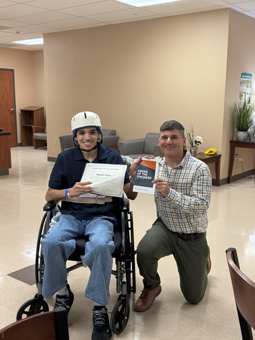 A man crouches next to another person in a wheelchair as they hold documents that read "Obligation of an Engineer" and "Order of the Engineer."