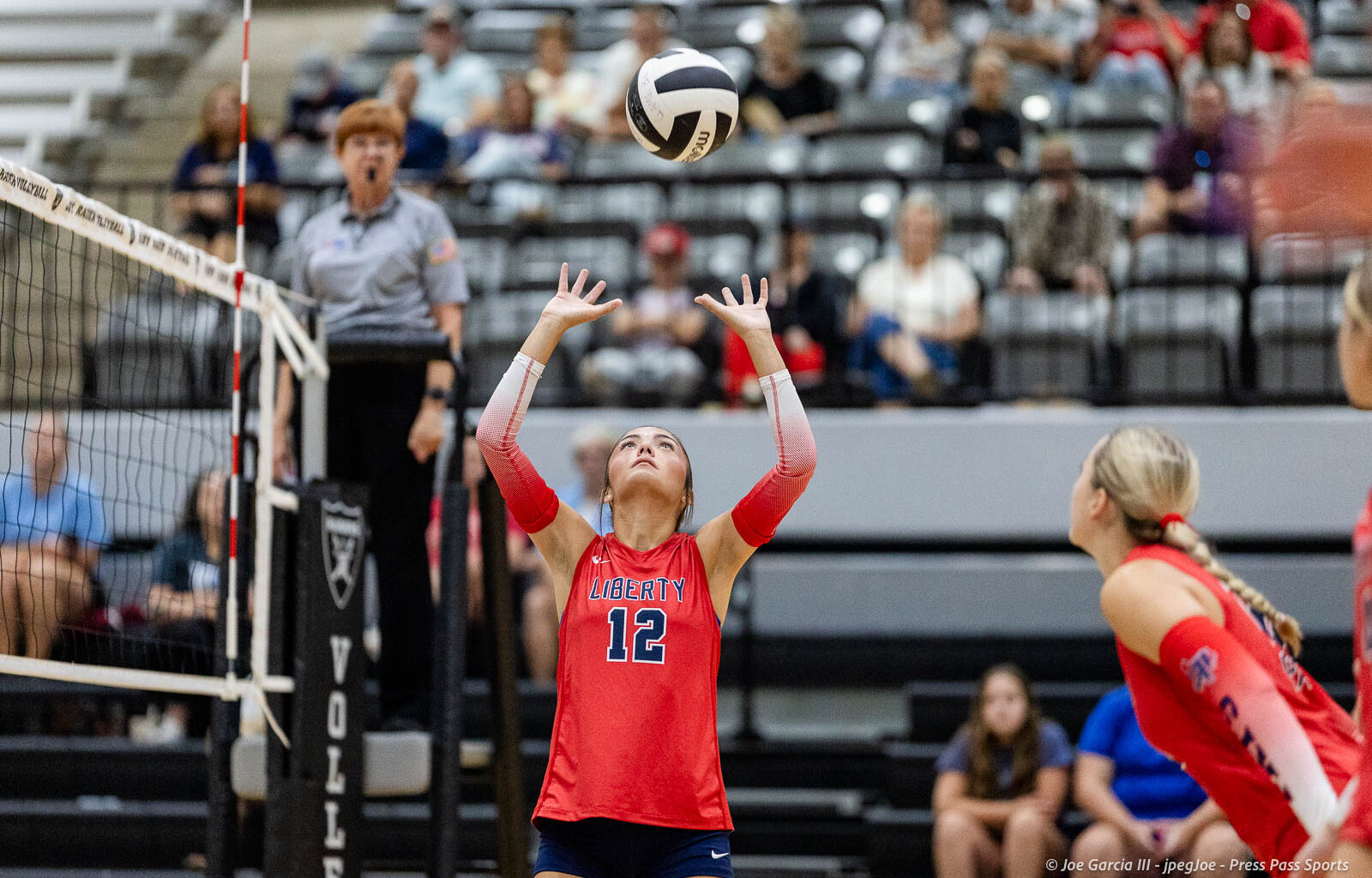 Liberty player #12 in red uniform raises hands to set the volleyball during a rally, with a referee and audience in the background.