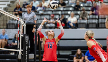 Liberty player #12 in red uniform raises hands to set the volleyball during a rally, with a referee and audience in the background.