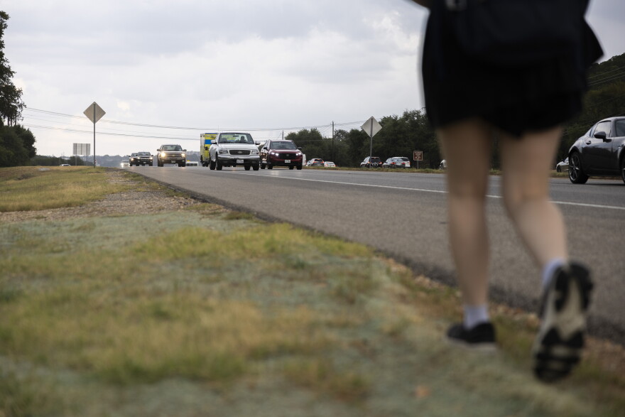 A person walks along a grassy shoulder beside a busy highway with multiple vehicles traveling in the opposite direction.