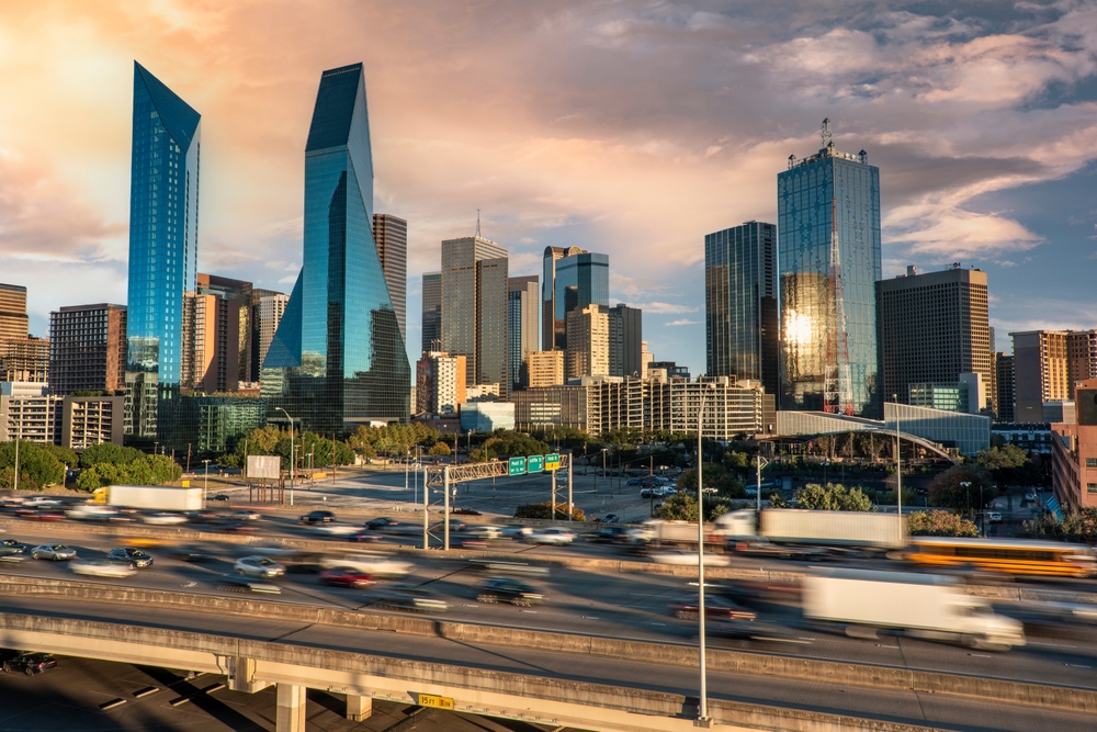 Vehicles travel on a freeway near downtown Dallas with the city skyline in the background