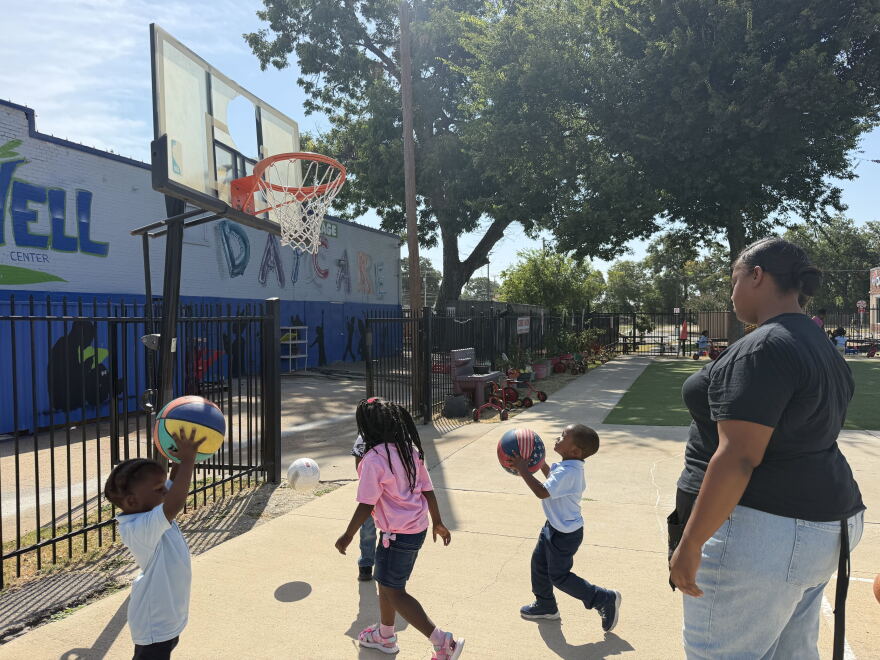 A woman in a black shirt and jeans watches as a group of pre-K students shoot basketballs at a hoop on an outdoor court.