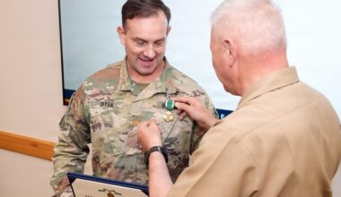 A military awards ceremony is taking place where a person in a tan/khaki uniform is pinning a medal onto a soldier in Army OCP (Operational Camouflage Pattern) uniform with the name tape
