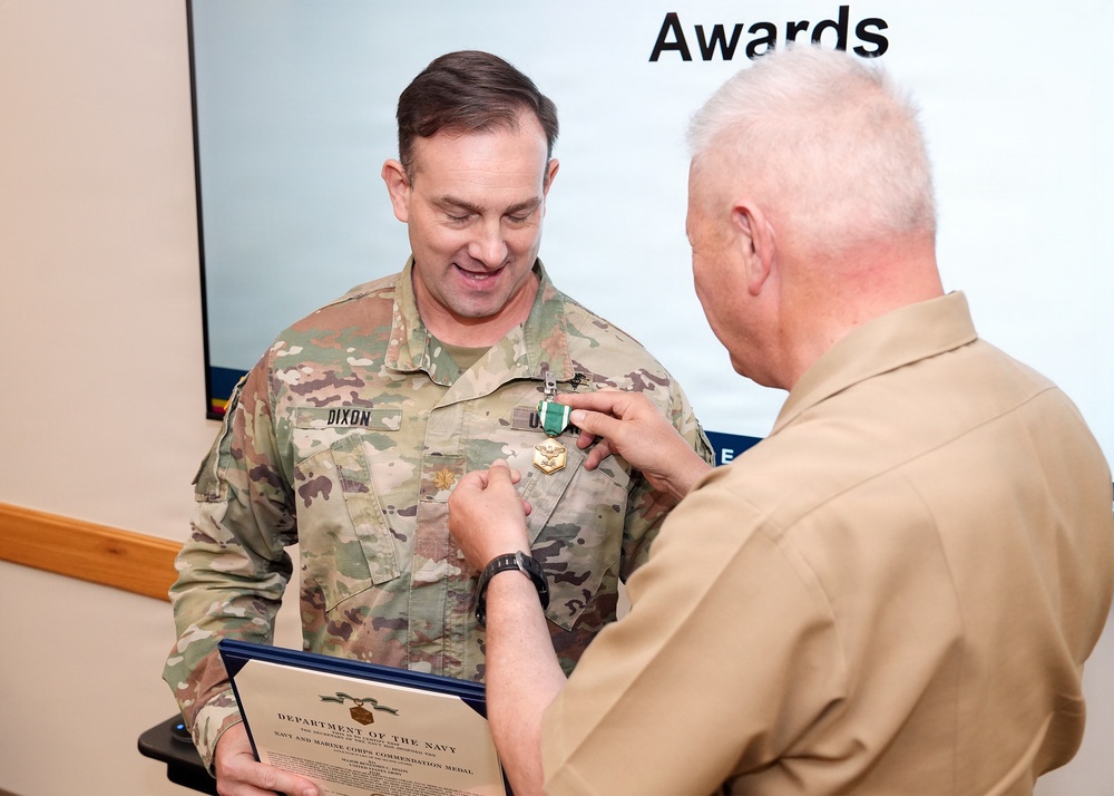 A military awards ceremony is taking place where a person in a tan/khaki uniform is pinning a medal onto a soldier in Army OCP (Operational Camouflage Pattern) uniform with the name tape