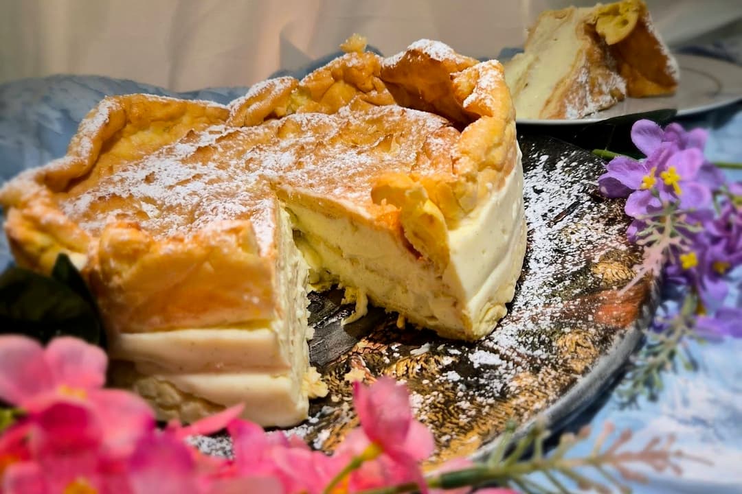 Cream-filled pastry cake dusted with powdered sugar, sliced open, surrounded by pink and purple flowers.