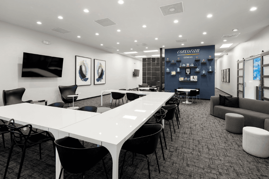 Modern conference room with white tables, black chairs, wall-mounted TV, and blue Corcoran Genesis branded accent wall.