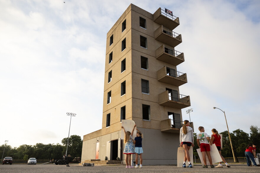 A group of children hold signs and cheer outside of a cement tower in a parking lot.
