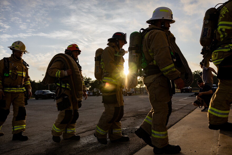 Partially silhouetted firefighters walk in front of a rising sun across pavement.