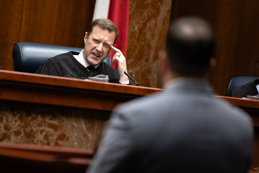 A judge in a black robe speaks into a microphone from the bench at a lawyer, whose back is visible in the foreground.