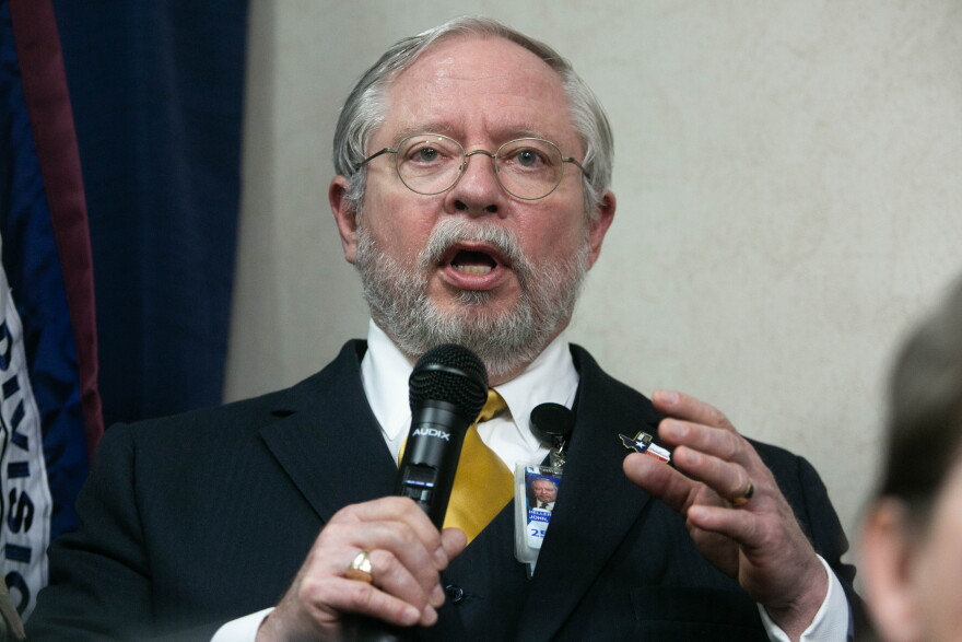 Former DSHS Commissioner John Hellerstedt wearing a suit and speaking into a handheld microphone.