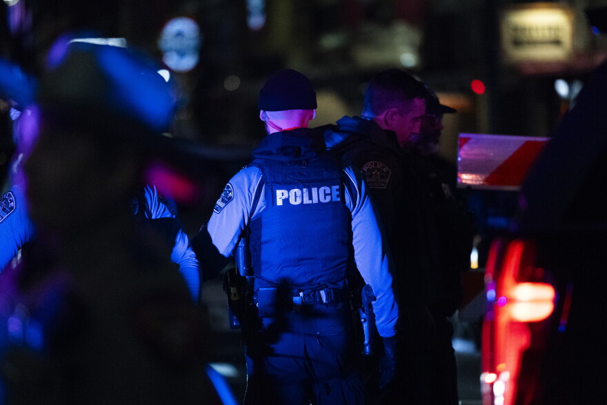 A group of police officers is gathered at night. Centered in the photo is the back of one officer, with the word "POLICE" printed on the back of a ballistic vest.