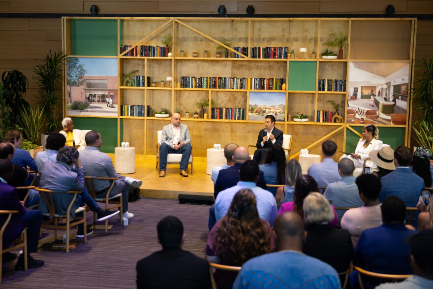 Four people sit in chairs with a background of bookshelves while speaking into microphones before a seated audience.