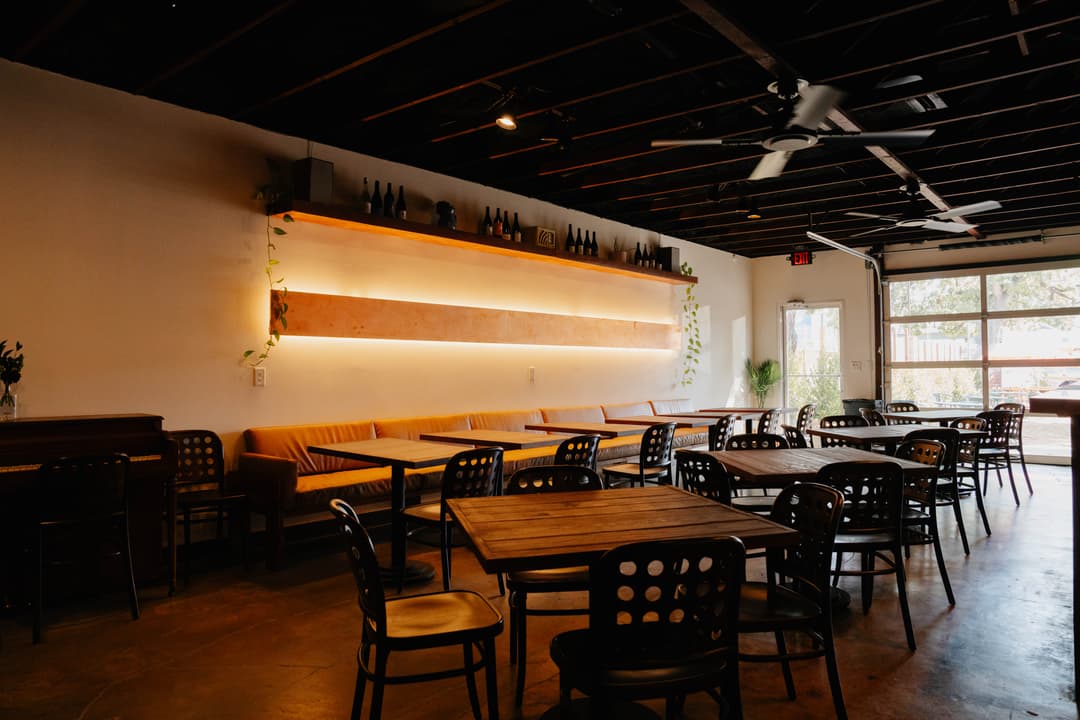 The image shows the interior of The Watershed with warm wood tables, booths, warm LED lighting and a garage door with open windows. 