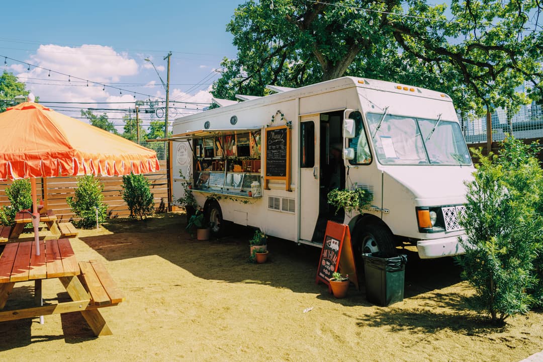 The image shows the exterior of the Muy Yum food truck, which is a white truck with pots and landscaping. The image also shows the picnic tables and orange umbrellas outside of The Watershed. 