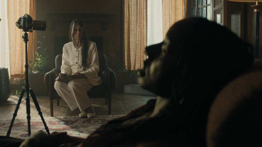 A still image from the short film Transference shows a therapist sitting in a chair talking with her patient. A camcorder is focused on the patient.