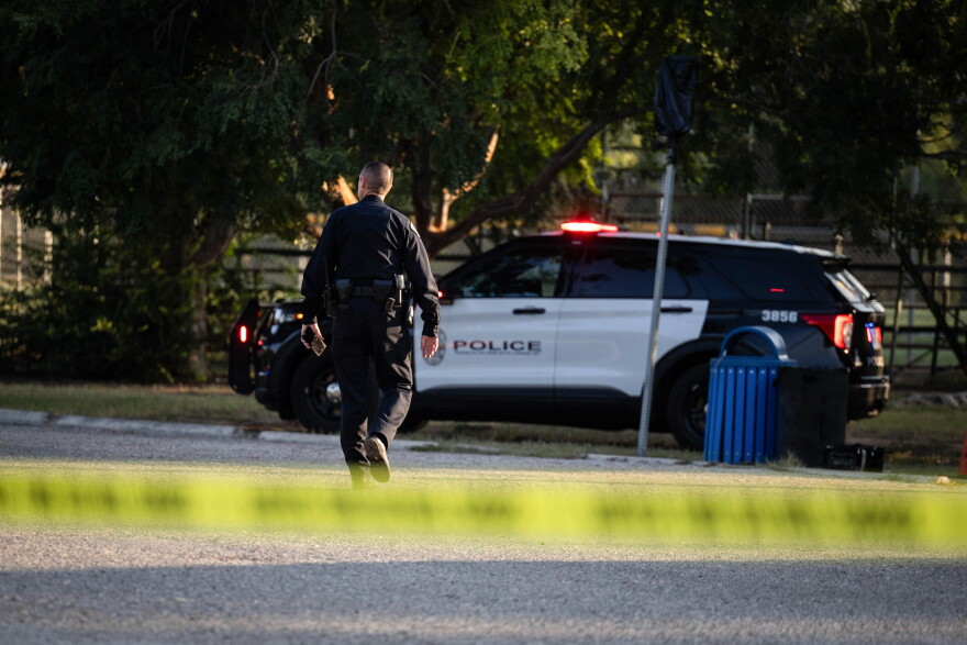 An Austin police officer walks toward a patrol vehicle. Yellow police tape is visible in the foreground.