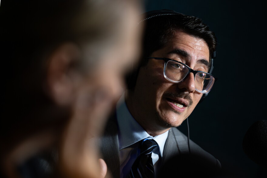 A man in a suit, wearing glasses, speaks while wearing headphones inside a KUT News radio studio.
