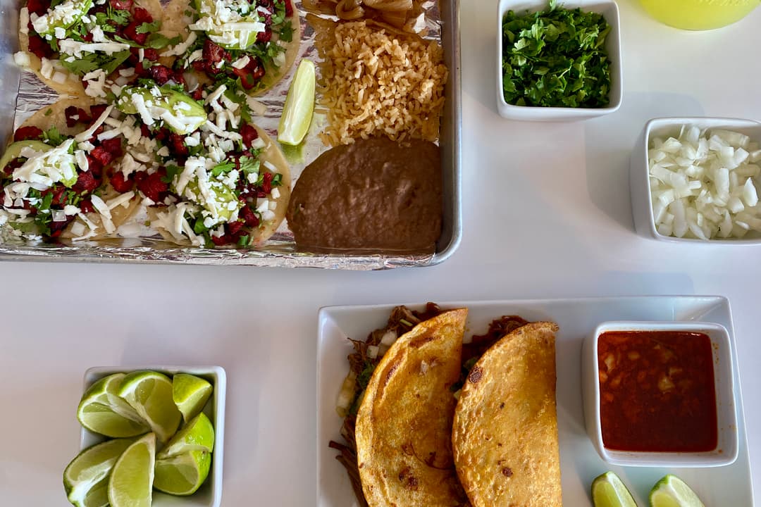 The image shows an a table with Mexican street-style tacos, queso birria tacos, onions, limes and salsas. 