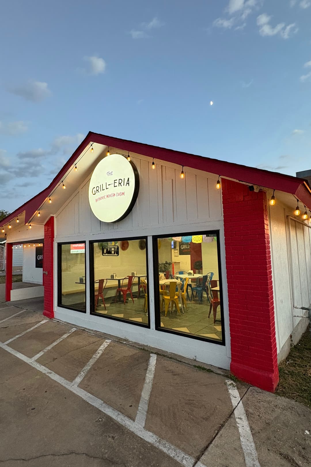 The image shows the exterior of Grill-eria which is a white building, red trim, string lights and a circular lit-up sign saying "Grill-eria."