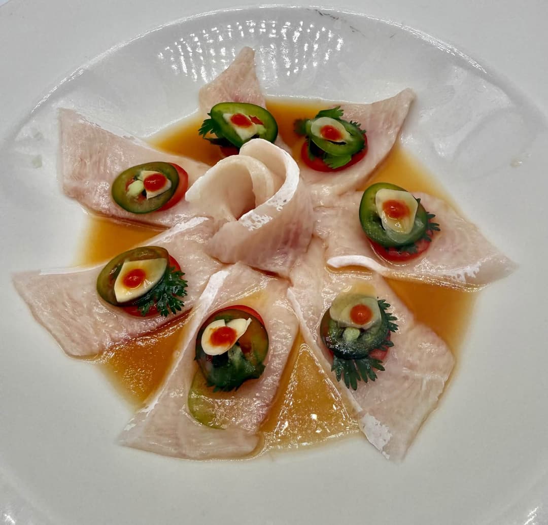 Yellowtail sashimi arranged in a circle, topped with jalapeño slices and hot sauce in citrus ponzu broth.