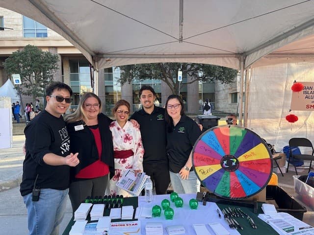 Five people pose behind an event booth with a colorful prize wheel, flyers, and giveaways under a tent.